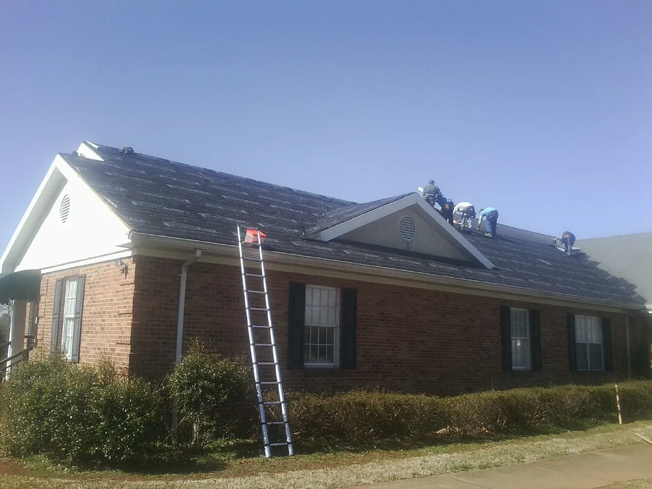 Crew tearing shingles off a brick ranch roof with a ladder set — Emergency Roof Repair work by our crew in Rich Hill