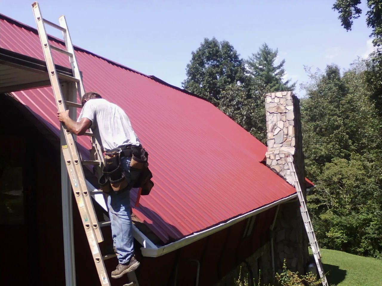 Roofer on a ladder beside a red metal roof and stone chimney — Soffit & Fascia Repair by Roofing Brothers in Rich Hill